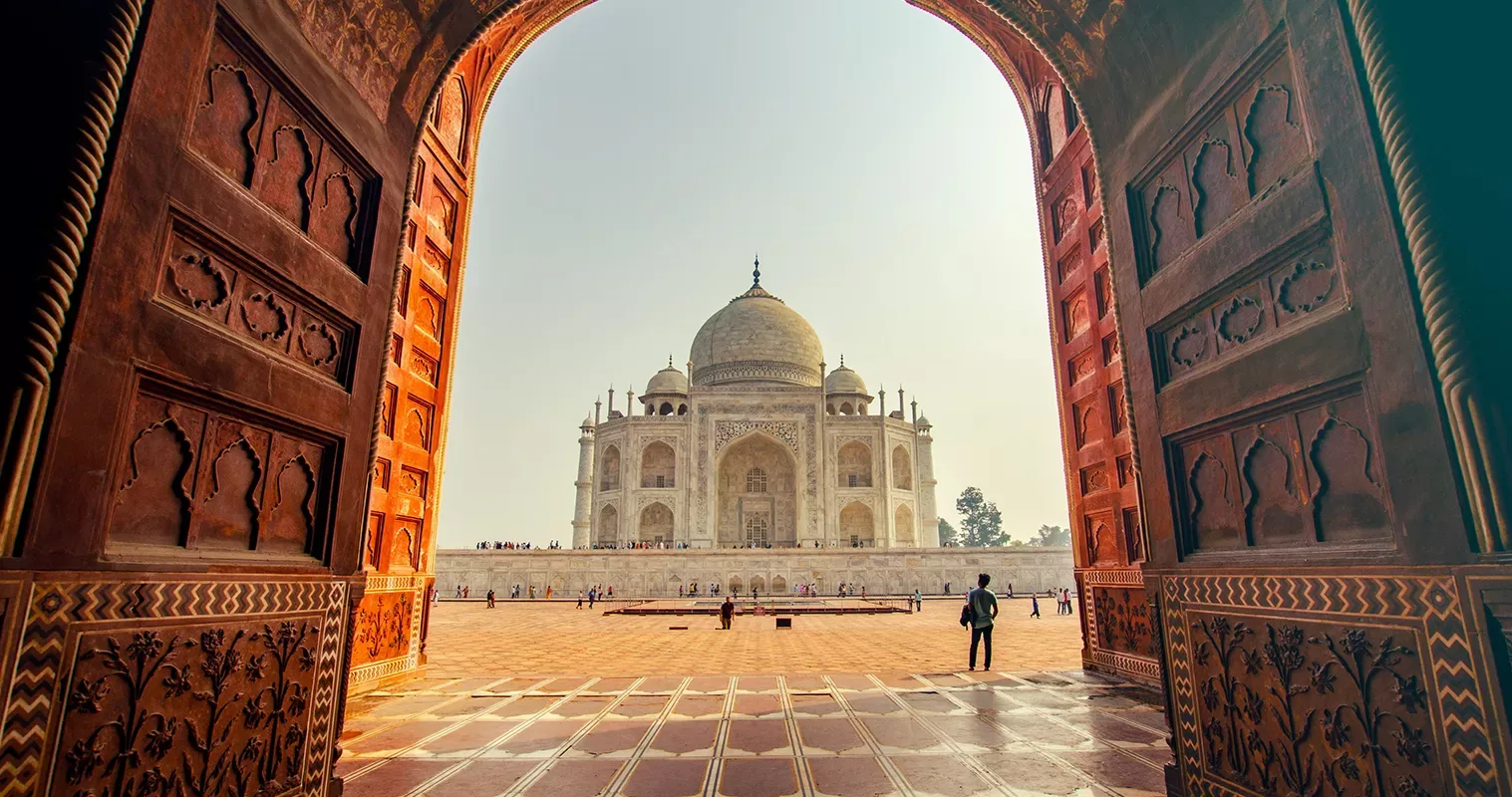 A view of the Taj Mahal framed by the large, ornate doorway of a grand entrance. Sunlight illuminates the white marble mausoleum against a clear sky, with a few visitors on the expansive red stone courtyard.