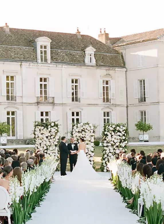 A couple stands at an altar adorned with white flowers, exchanging vows in front of a historic mansion. Rows of guests are seated on either side of a flower-lined aisle under a clear sky.