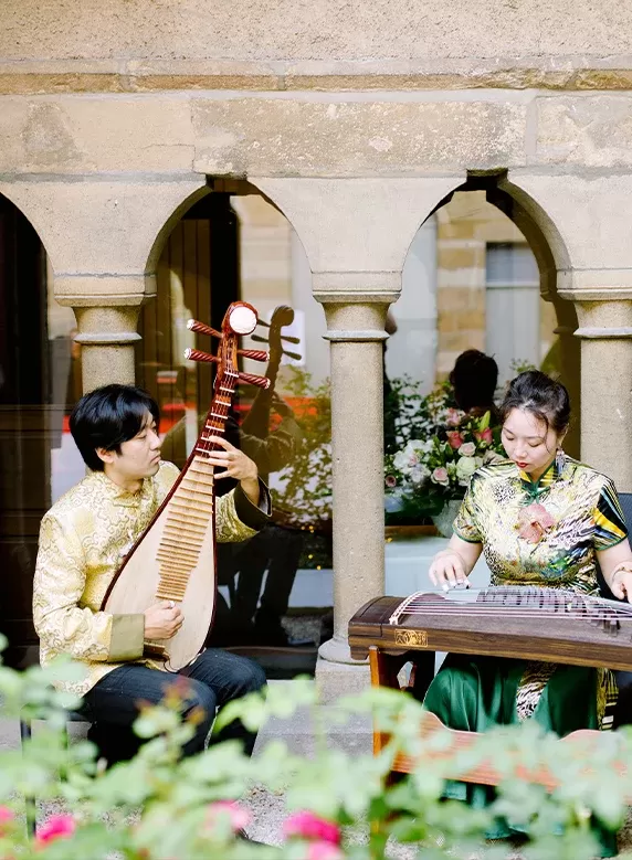 Two musicians in traditional attire perform in a stone archway. One plays a pipa, and the other plays a guzheng. Flowers and greenery are visible in the foreground, with a window reflecting more flowers in the background.
