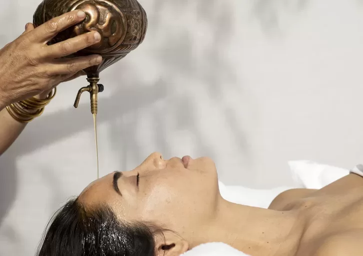 A person lies on their back with eyes closed as oil is gently poured onto their forehead from a brass vessel, suggesting a relaxing spa or Ayurvedic treatment.
