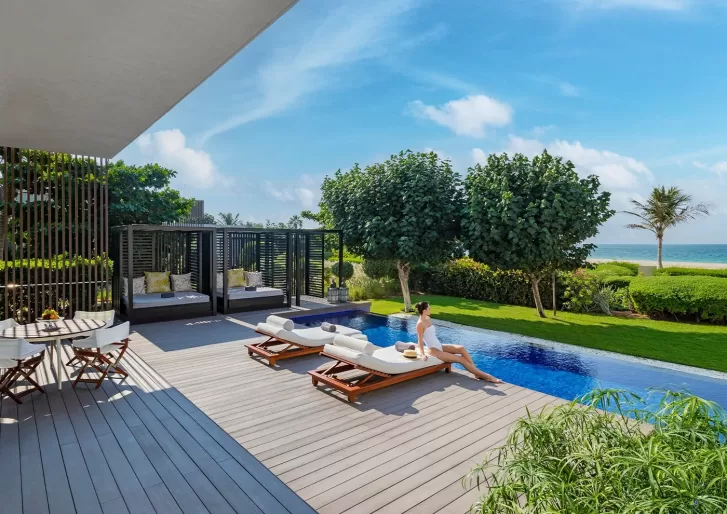 A woman relaxes on a lounge chair by an outdoor pool overlooking the ocean. The scene includes lush greenery, modern cabanas, and a small dining table on a wooden deck under a clear blue sky.