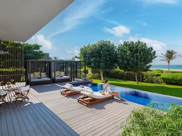 A woman relaxes on a lounge chair by an outdoor pool overlooking the ocean. The scene includes lush greenery, modern cabanas, and a small dining table on a wooden deck under a clear blue sky.