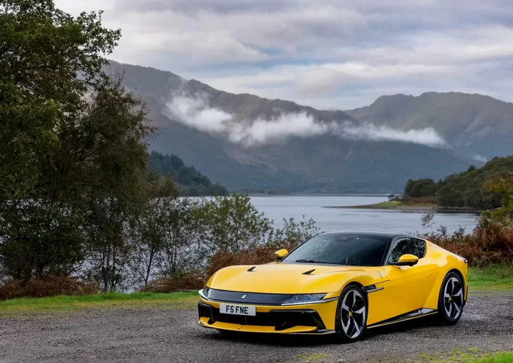 A yellow sports car parked on a dirt road by a lake, with green trees and mountains in the background under a cloudy sky.