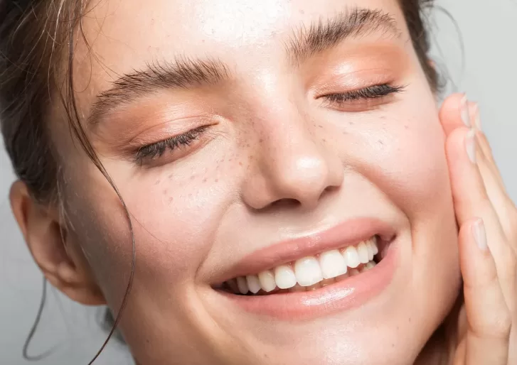 A person with closed eyes and a big smile touches their cheek with one hand, showcasing clear skin and natural makeup with soft, peach-colored eyeshadow.