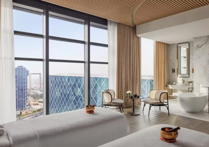 A beautiful room with floor to ceiling windows overlooks Dubai. Inside the room is 2 white massage beds with a golden sound bowl placed on each bed.