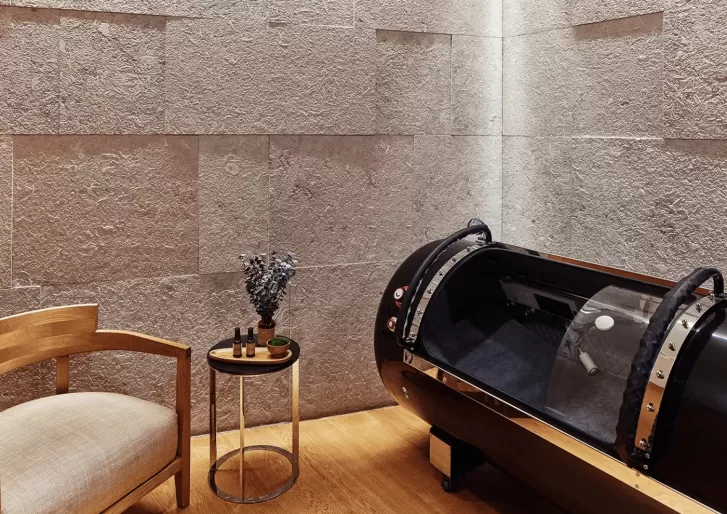 A minimalist room featuring a wooden chair with a beige cushion, a small round table with artful decorations, and a sleek black pod-like structure, all set against textured stone walls at Longevity at Bvlgari Spa.