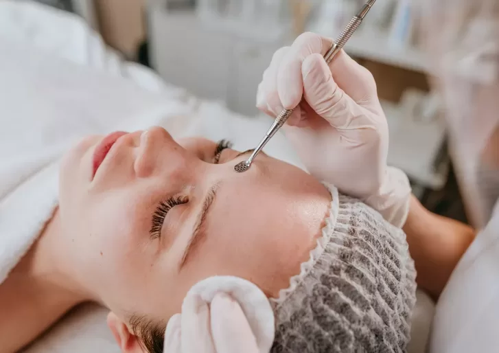 A person is lying on a treatment bed, wearing a hair net. A gloved professional is using a metal tool on their forehead during a skincare procedure. A cotton pad is placed on the side of the persons face.