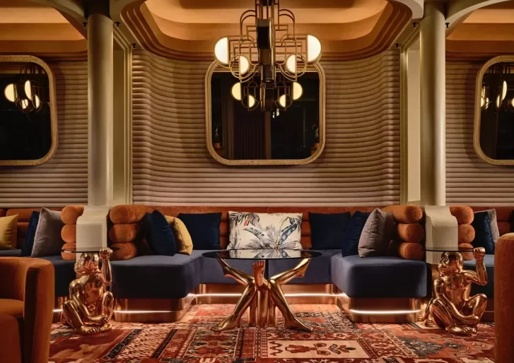 Stylish lounge with a luxurious interior featuring a navy blue sectional sofa, ornate chandelier, geometric wall patterns, a glass coffee table with a star-shaped gold base, and gold dog sculptures. Richly patterned rug covers the floor.