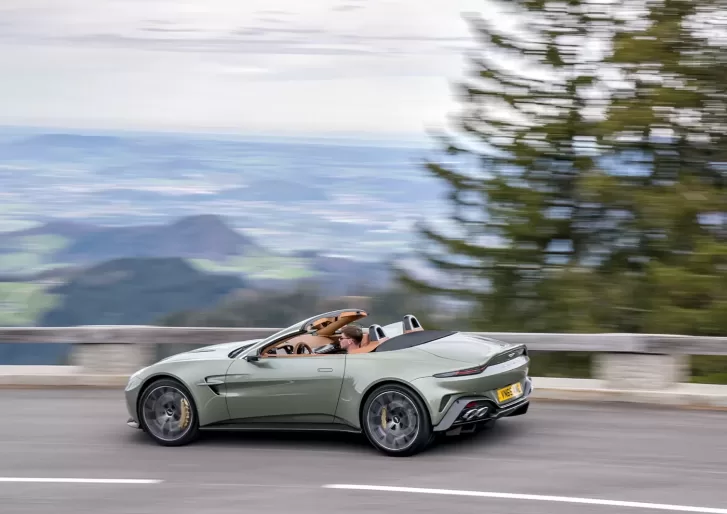 A light green convertible sports car with the top down drives quickly along a mountain road, passing trees with a scenic valley visible in the background.