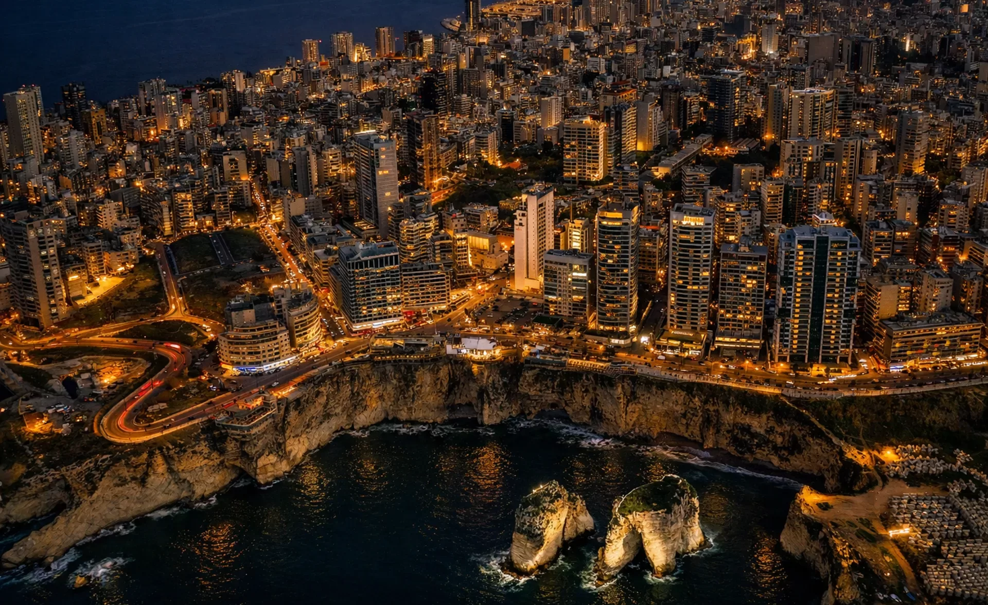 Aerial view of a coastal city at dusk with illuminated buildings, busy streets, rocky cliffs, and two large sea stacks in the water below.