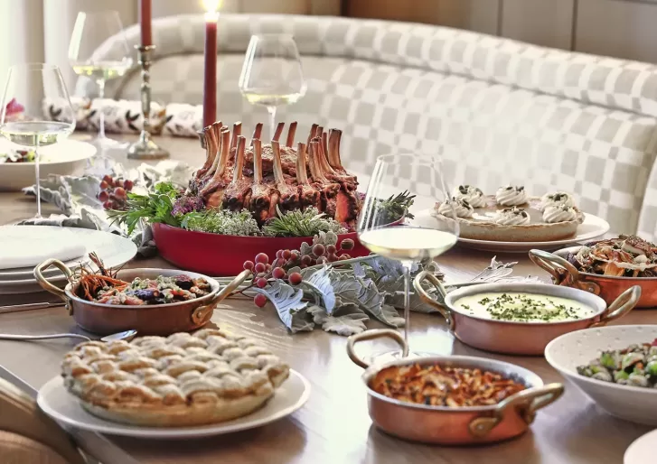A festive dining table set with wine glasses, candles, and various dishes including a roasted rack of lamb, sides in copper pans, a pie, salads, and pavlova desserts, decorated with fresh greens and berries.