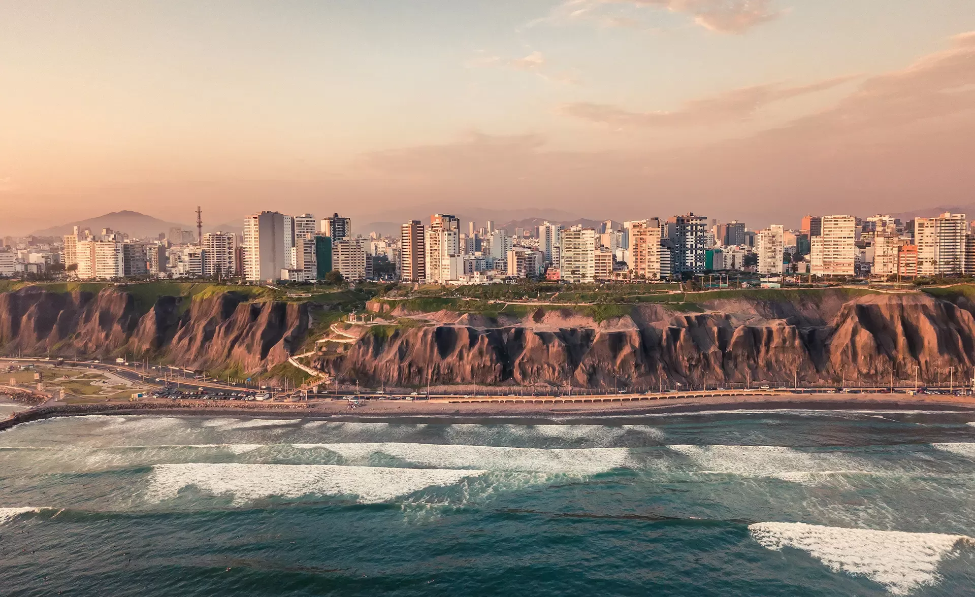 Aerial view of a coastal cityscape with tall buildings lined along cliffs overlooking the ocean. Waves crash against the shore under a sky painted with warm, pastel hues of sunset, creating a serene and picturesque scene.