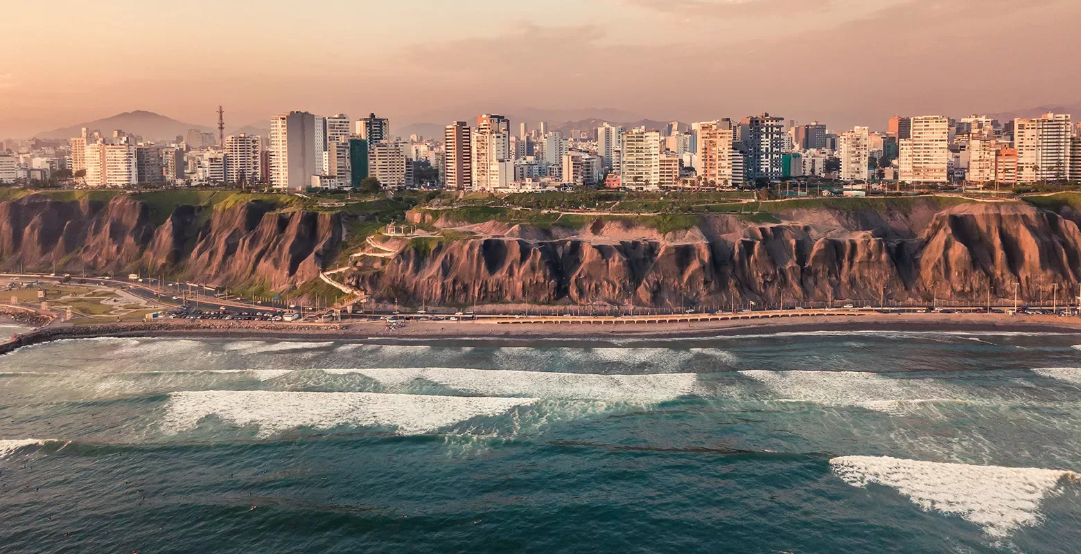 Aerial view of a coastal city with modern skyscrapers lining the cliffside. Waves crash onto the shoreline below, and a mixture of greenery and urban development is visible under a warm, hazy sky.