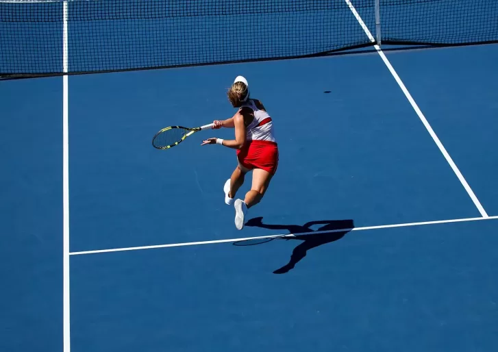 A tennis player in a red skirt and white top jumps to hit a forehand shot on a blue hardcourt, with the net and white boundary lines visible. The players shadow is cast on the court.