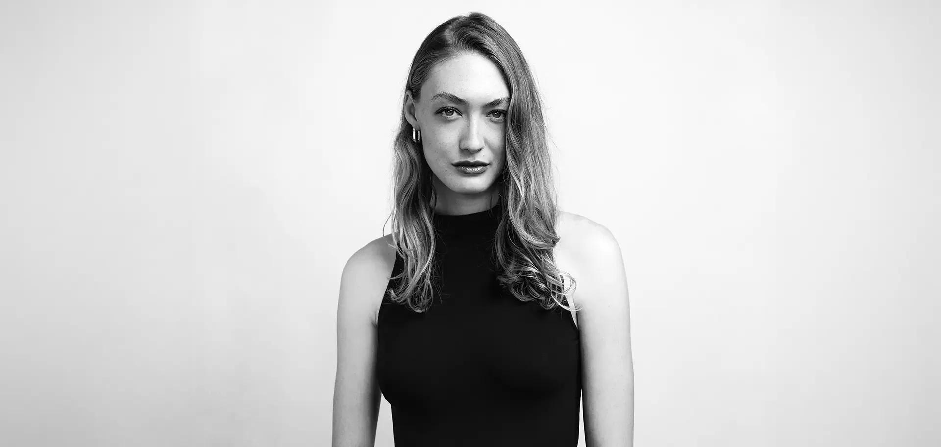 A woman with long, wavy hair wearing a sleeveless black top stands against a plain, light-colored background, looking directly at the camera with a neutral expression. The image is in black and white.