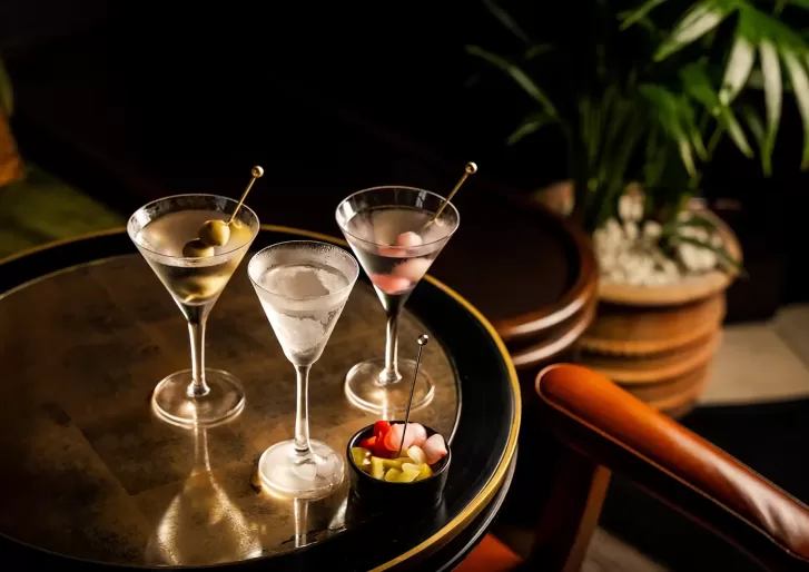 Three martini glasses with olives and cocktail onions are arranged on a round, reflective table next to a small bowl of garnishes. There is an orange chair and a potted plant in the background.