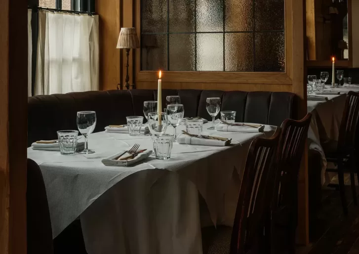 A dimly lit restaurant table set for four with white tablecloth, glassware, plates, cutlery, and a single lit candle, surrounded by dark wooden chairs and a cushioned booth.