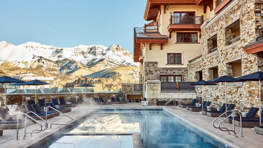 Outdoor heated pool surrounded by lounge chairs and umbrellas at a stone and wood mountain resort, with snow-capped peaks and blue sky in the background. Steam rises from the pool in the crisp air.