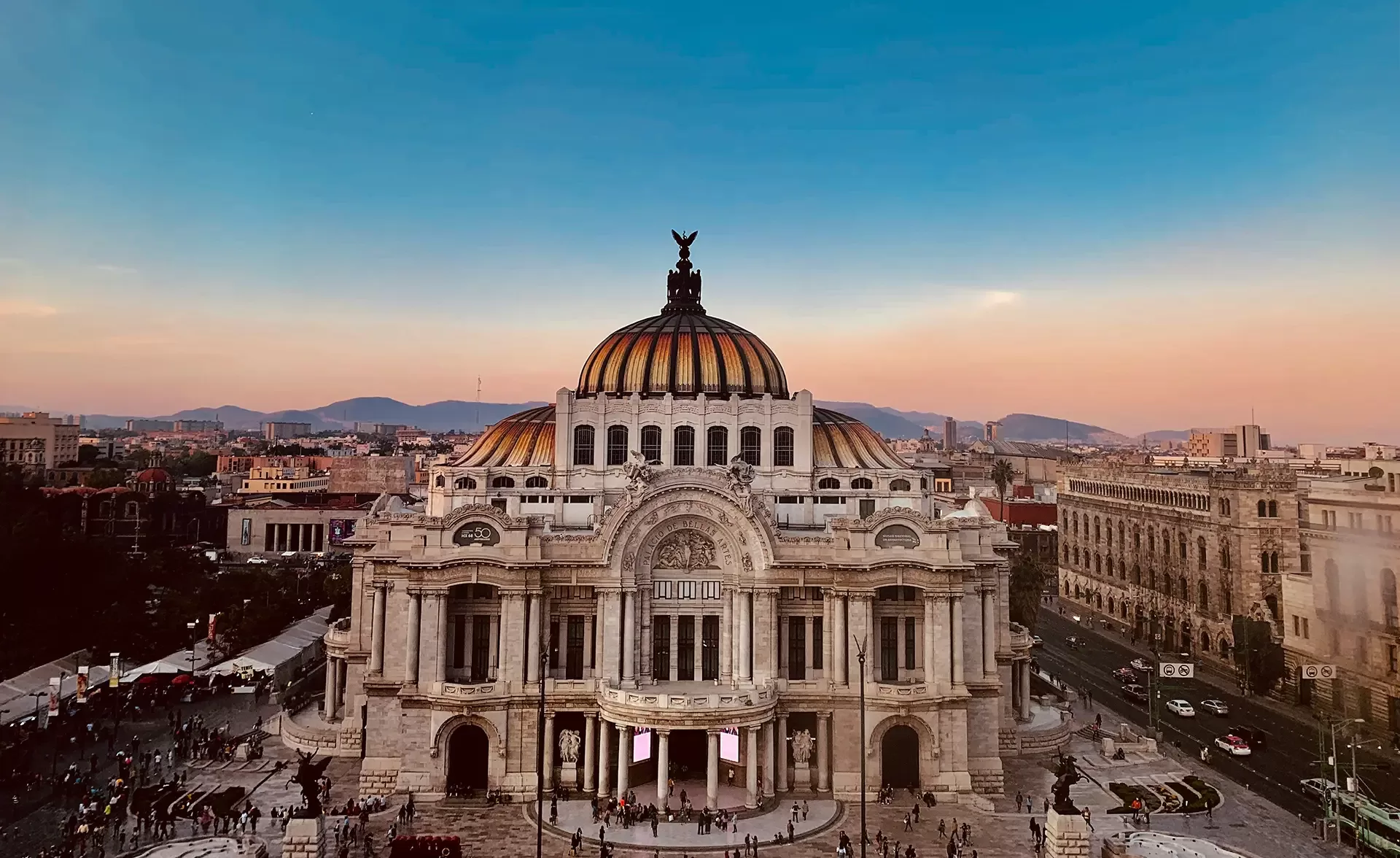 A grand, ornate building with a domed roof stands at sunset, surrounded by a bustling cityscape. The sky fades from blue to warm hues, with mountains in the distance. Crowds gather near the entrance, highlighting the buildings architectural beauty.