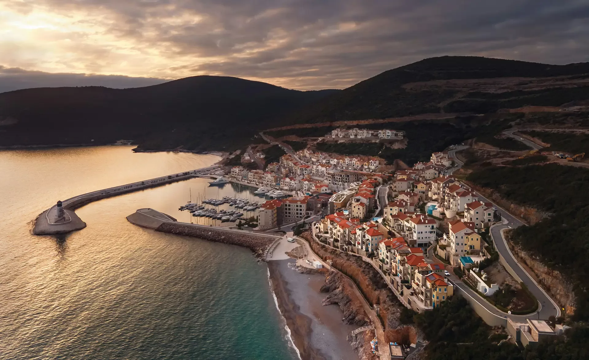 Aerial view of a coastal town at sunset, featuring a harbor with moored boats, buildings with red-tiled roofs, and surrounding green hills. The sky is partly cloudy, casting a warm glow over the calm sea and shoreline.