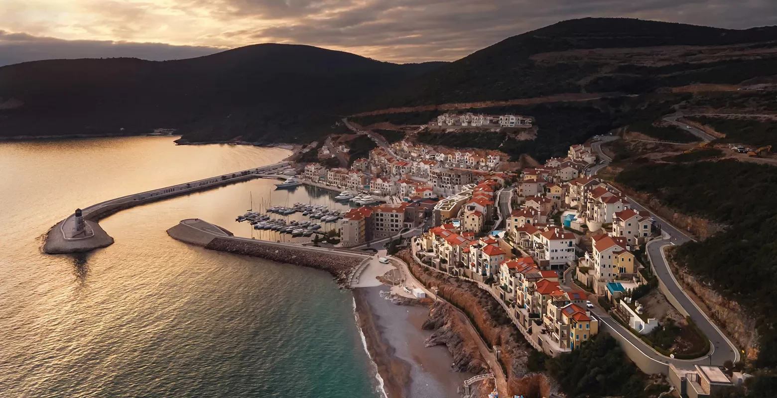 A coastal town at sunset with red-roofed buildings nestled between hills and the sea. A winding road leads through the town, and a harbor with boats is nestled along the shoreline. The sky is dramatic with dark clouds and golden light.