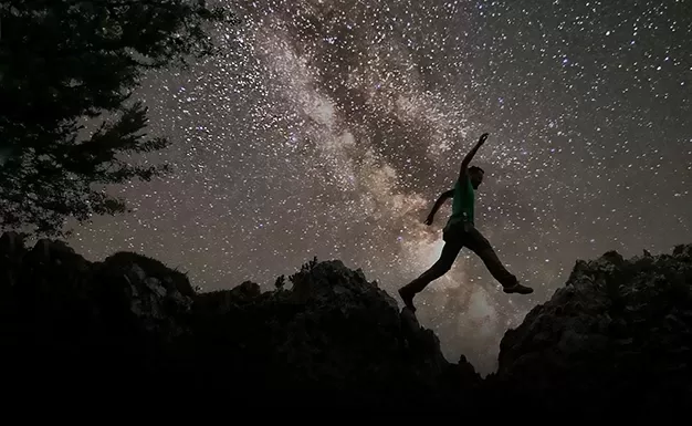 Silhouette of a person leaping between two rocky outcrops under a starry night sky, with the Milky Way galaxy visible in the background. A tree is partially visible on the left.