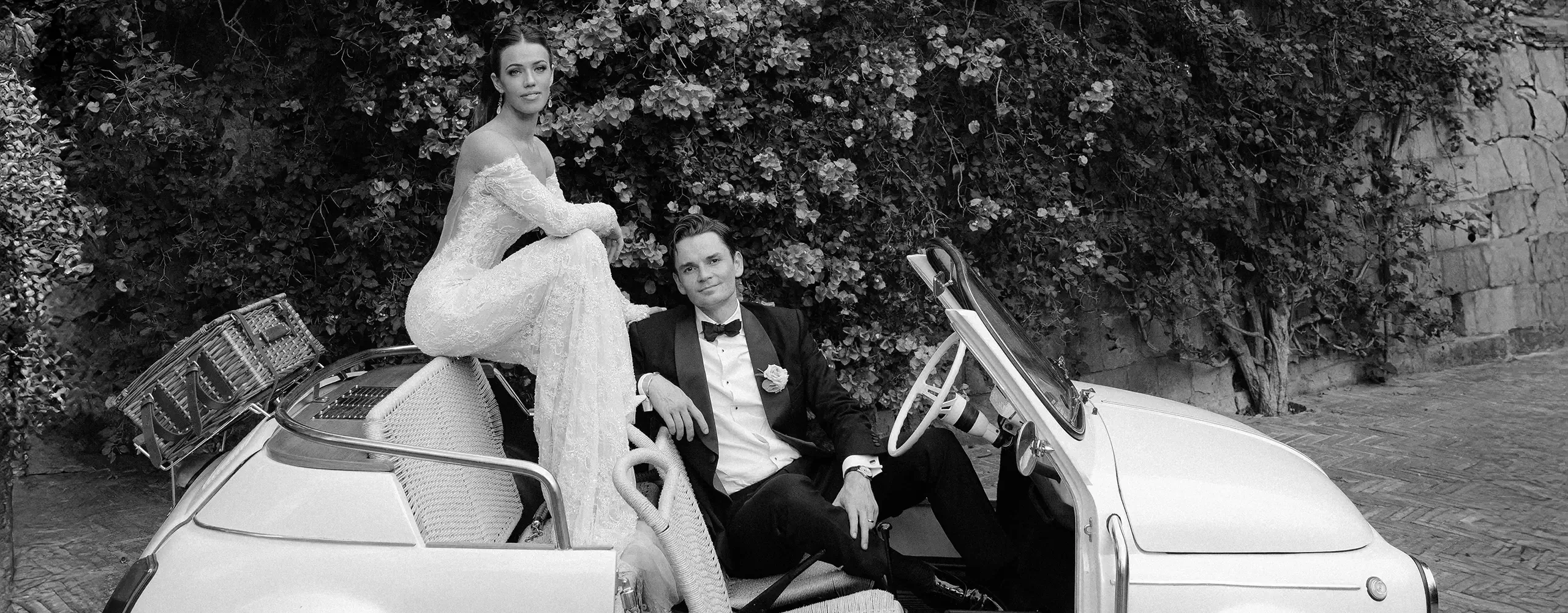 A bride in an off-the-shoulder gown sits on the back of a vintage convertible, while a groom in a tuxedo sits in the driver’s seat; they pose together in front of a leafy wall, looking at the camera.