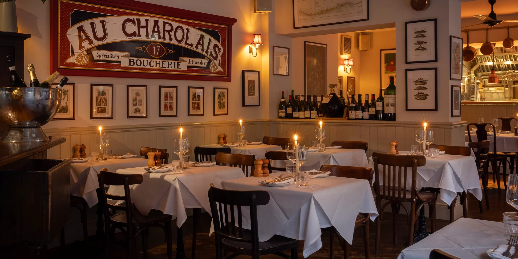 A cosy French restaurant with white tablecloths, wooden chairs, lit candles on tables, wine bottles on a shelf, framed art on the walls, and a view into the kitchen in the background.