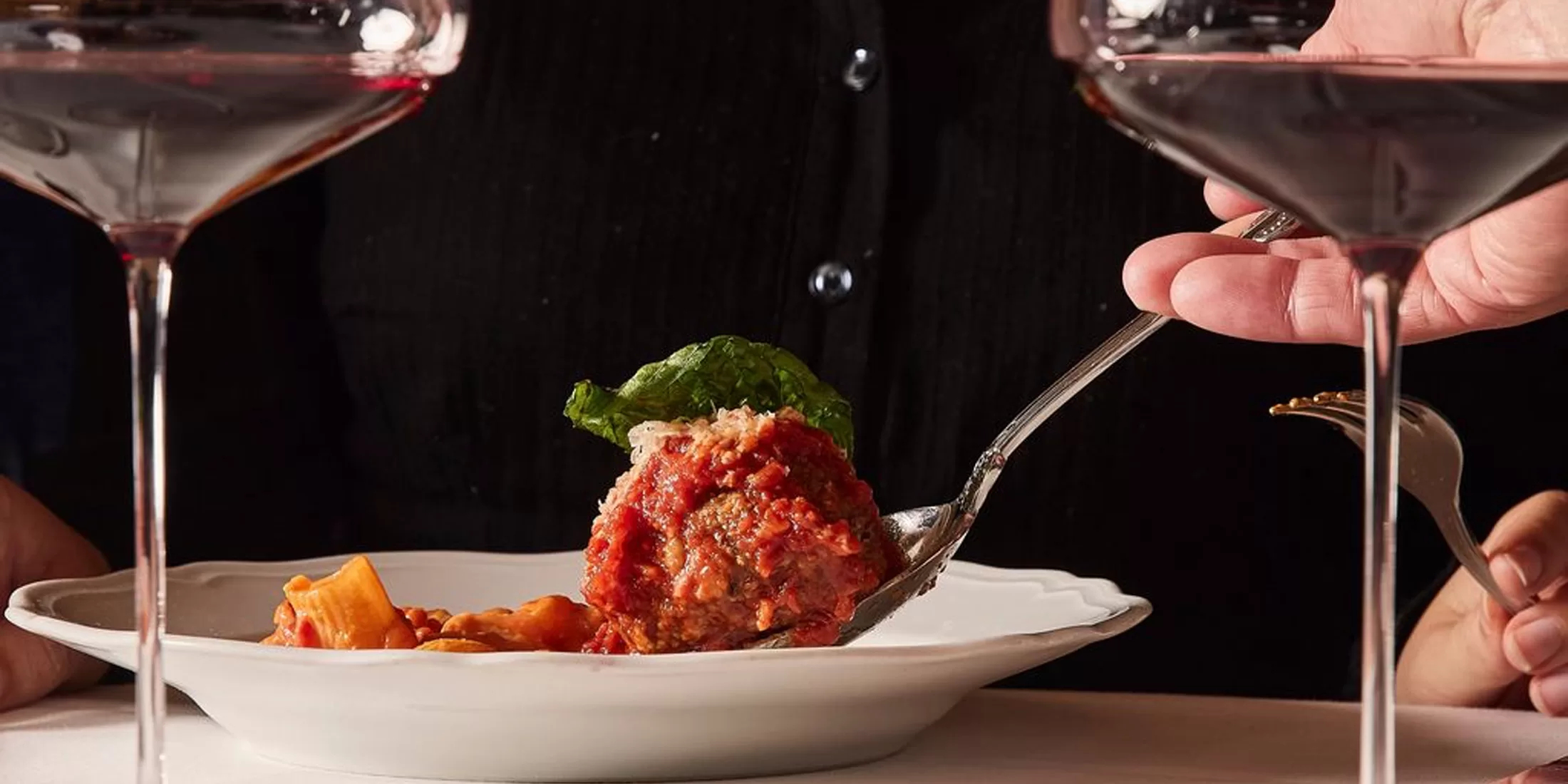 A person serves a large meatball with tomato sauce and pasta onto a white plate, surrounded by two glasses of red wine on a table.
