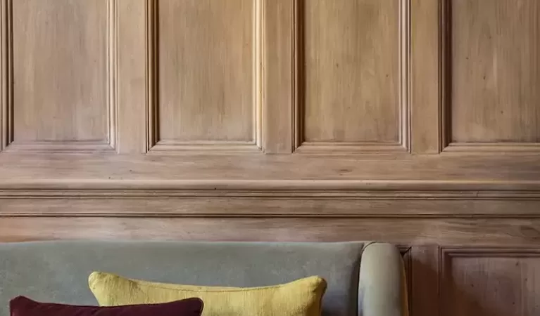 A round wooden table set with three glasses of water, three white napkins, and cushioned seating. The background features paneled wood walls and a wall sconce with a frosted glass shade.