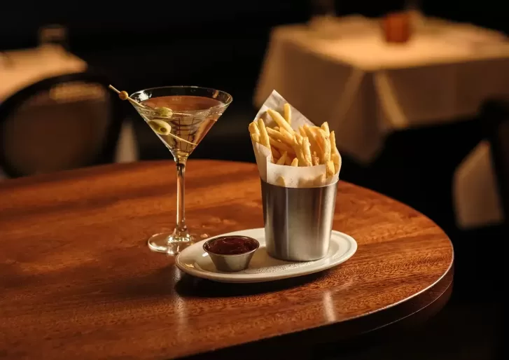 A glass of martini with olives and a metal cup filled with French fries, served with ketchup on a small white plate, sitting on a wooden table in a dimly lit restaurant.
