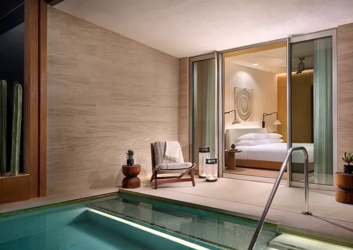 A modern hotel room with sliding glass doors opens to a private patio featuring a chair, small tables with plants, and a pool with a metal handrail. The room has neutral tones and contemporary decor.