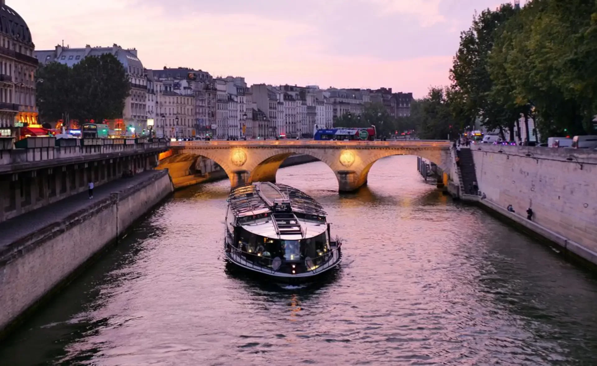 A boat cruises along the Seine River at sunset in Paris, passing under a stone bridge, with historic buildings and glowing streetlights lining the riverside.