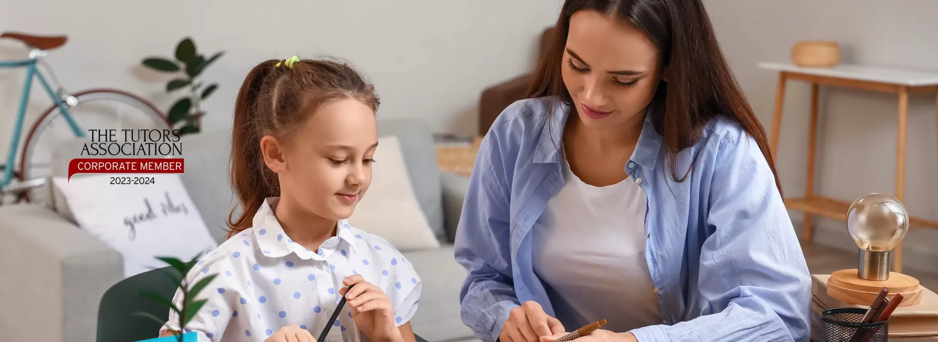 A woman and a young girl sit at a table working on schoolwork together, both smiling. A logo says The Tutors Association Corporate Member 2023/2024 on the left. A plant and a bicycle are in the background.