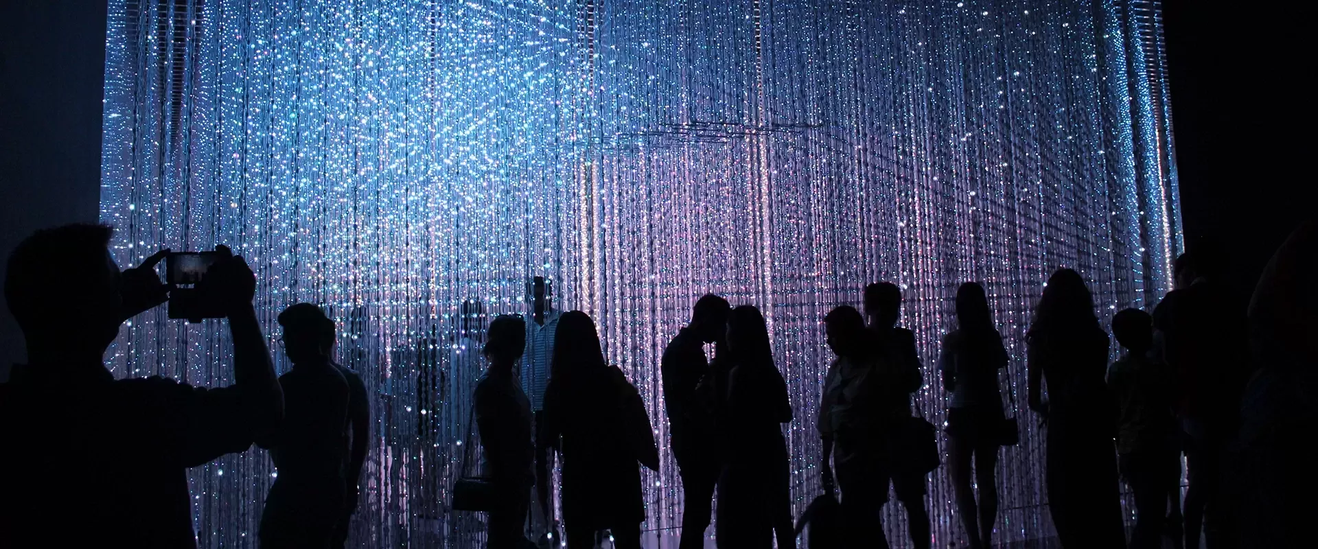 Black silhouettes of people walking through and taking pictures of a light exhibition in a gallery