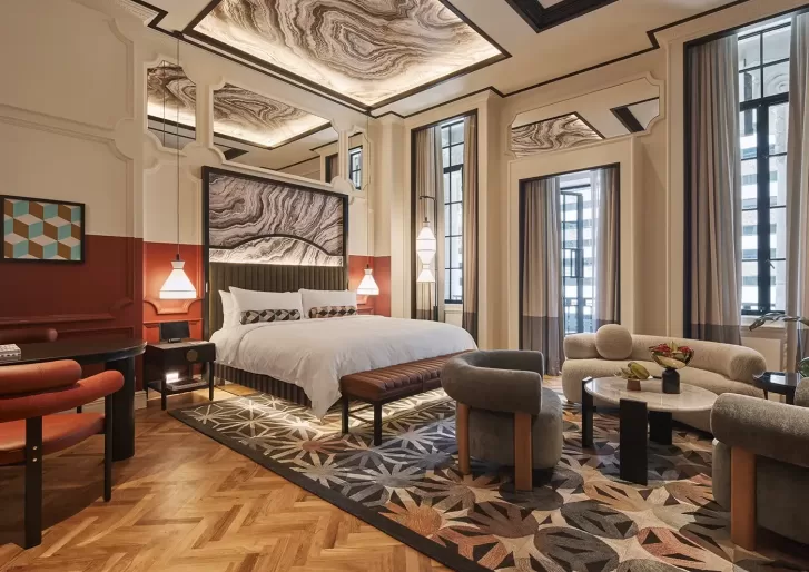 A luxurious hotel room with a large bed, geometric-patterned carpet, and modern furnishings. The ceiling features decorative panels, and the room is illuminated by stylish pendant lights and large windows.