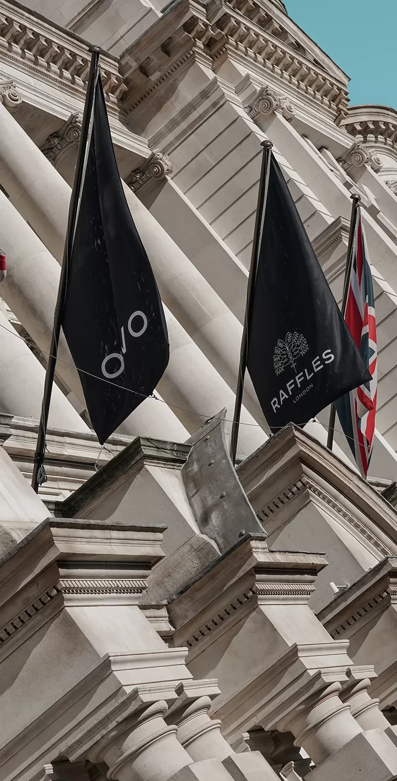 Three flags hang from a stone building: a black OVO flag, a black Raffles London flag, and a British flag. The detailed building facade features columns and ornate architectural details under a clear blue sky.