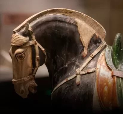 Close-up of an ancient terracotta horse sculpture, featuring intricate details in the mane, bridle, and saddle. The horses head is tilted gracefully, capturing a realistic yet artistic essence. The background is blurred, focusing attention on the sculpture.