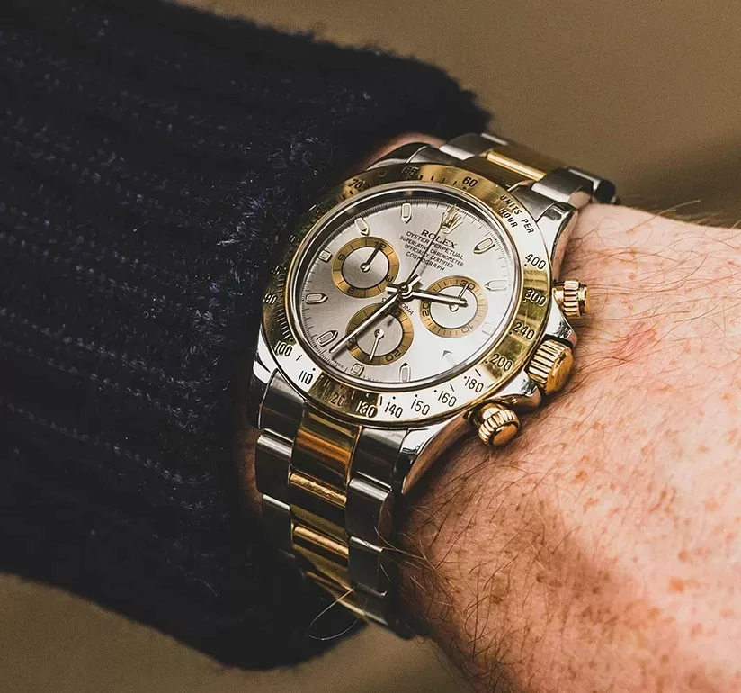 A silver and gold wristwatch with three subdials and a tachymetric scale on the bezel, worn on a persons wrist. The watch features a mix of metallic links on the band and a dark sweater sleeve partially visible.