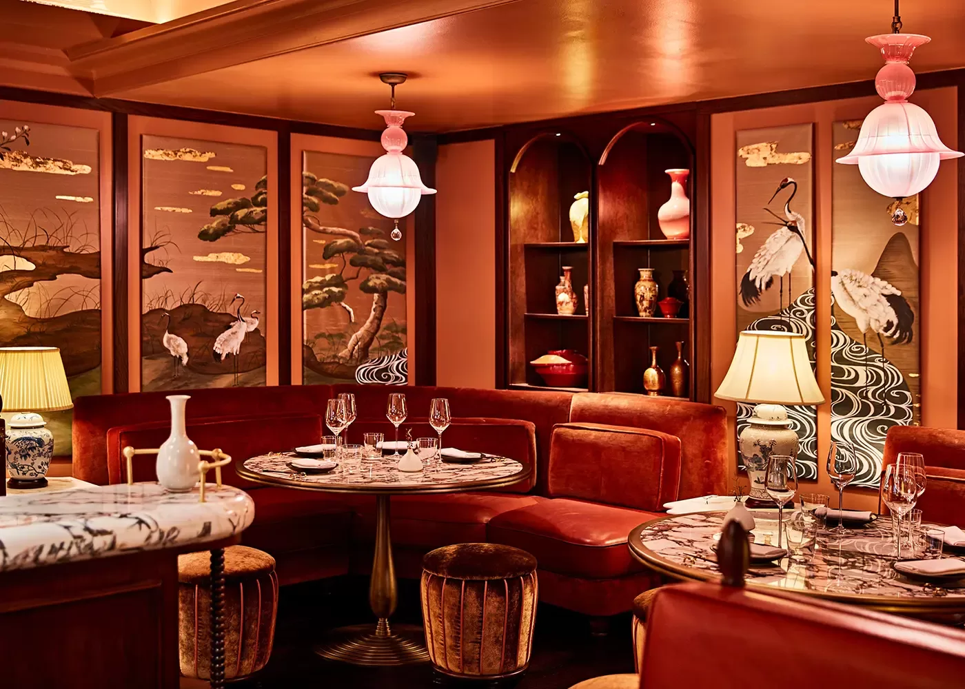 Elegant restaurant interior with round marble tables, plush red seating, and decorative screens featuring cranes and trees. Warm lighting from lamps and pendant lights, with shelves displaying vases and ceramics.