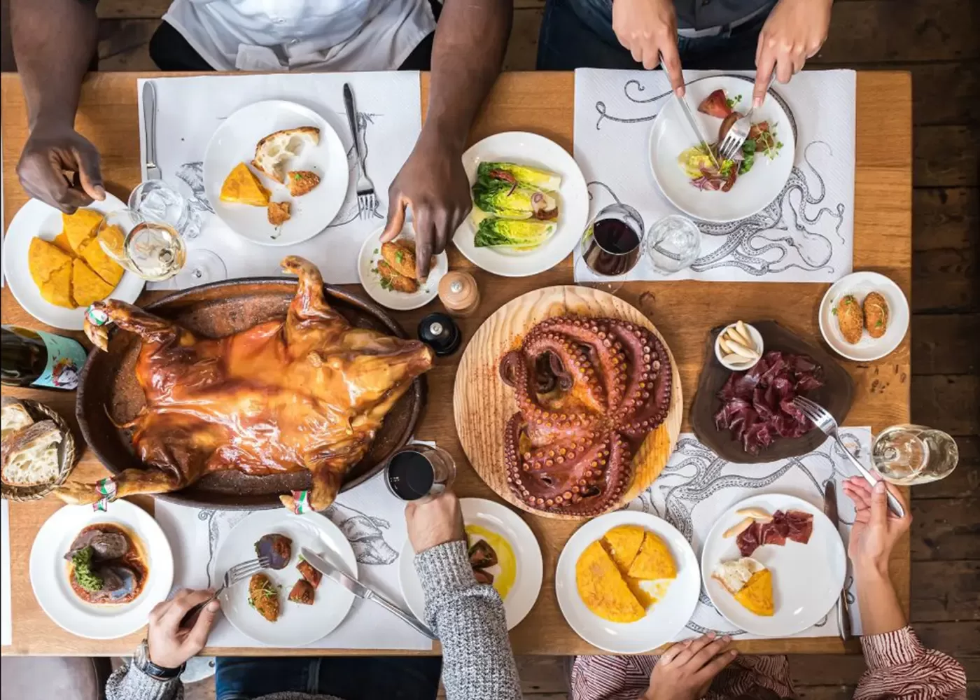 A table set for four with a variety of dishes, including a whole roast pig, a large plate of cooked octopus, salads, bread, and glasses of red wine. Four people are seated, sharing and eating the meal.