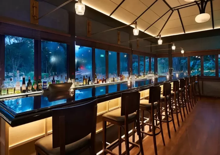 A modern bar with high wooden chairs lined along a glossy counter, shelves holding various bottles, and large windows revealing a nighttime outdoor view with trees and blue lighting. Warm ceiling lights illuminate the space.