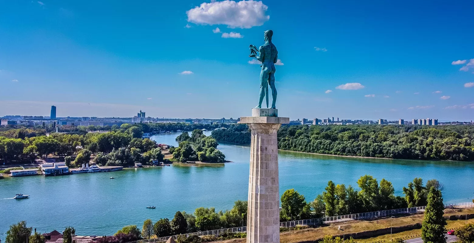 A tall statue stands on a pedestal overlooking the confluence of two rivers, surrounded by lush greenery and a distant urban skyline under a clear blue sky with a few clouds.