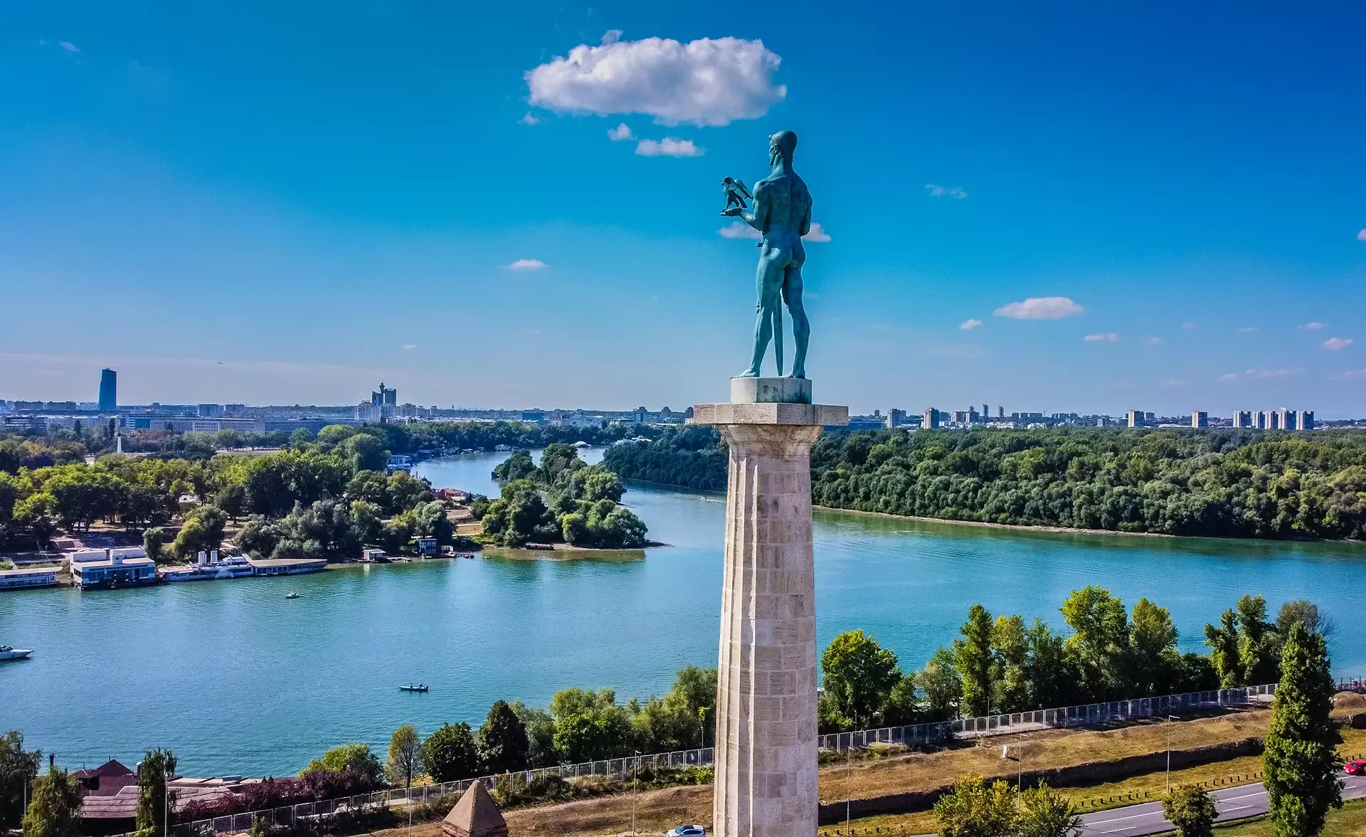 A tall statue of a man holding a bird and a sword stands atop a pillar overlooking a wide river with a scenic city and forest landscape. A few clouds dot the bright blue sky, and the river is lined with green trees.