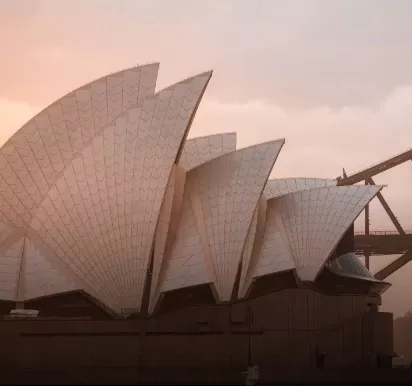 The image shows the Sydney Opera House with its iconic white sail-like structures at sunset. The sky is a warm gradient, and part of the Sydney Harbour Bridge is visible in the background.
