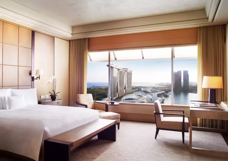 Hotel room with a double bed, armchair, desk, and lamp. Large window offers a view of iconic buildings and a body of water. Neutral tones and wood accents create a cozy atmosphere.