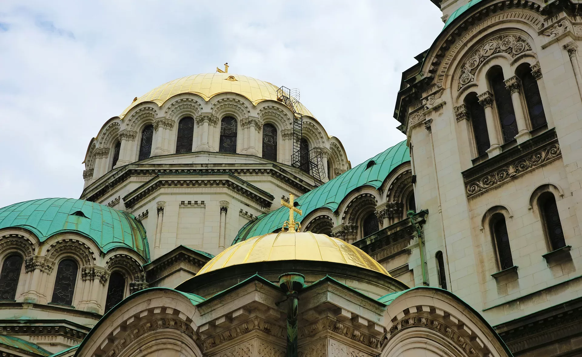 A historical building with ornate architecture featuring multiple domes; some are golden and others are patina green. The sky is cloudy, adding a dramatic backdrop to the intricate details and arched windows of the structure.