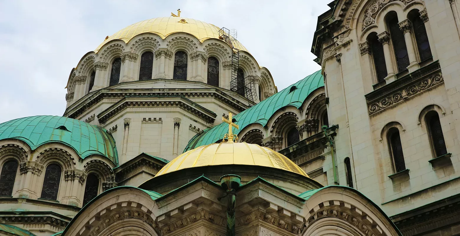 Close-up of a historic church with a blend of green and gold domes. The architecture features ornate arches and detailed stonework. The sky is overcast, highlighting the intricate design of the building.