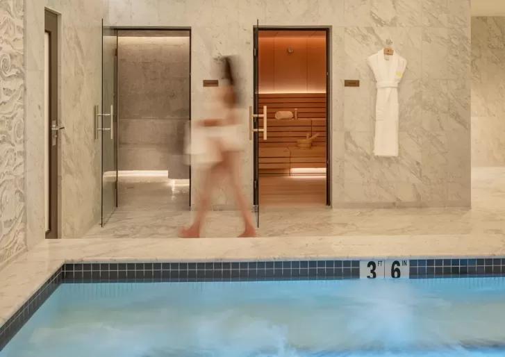 A blurred person in a towel walks past a sauna and steam room in a marble spa area, with a robe hanging on the wall and a pool with bubbling water in the foreground marked 3 ft 6 in.
