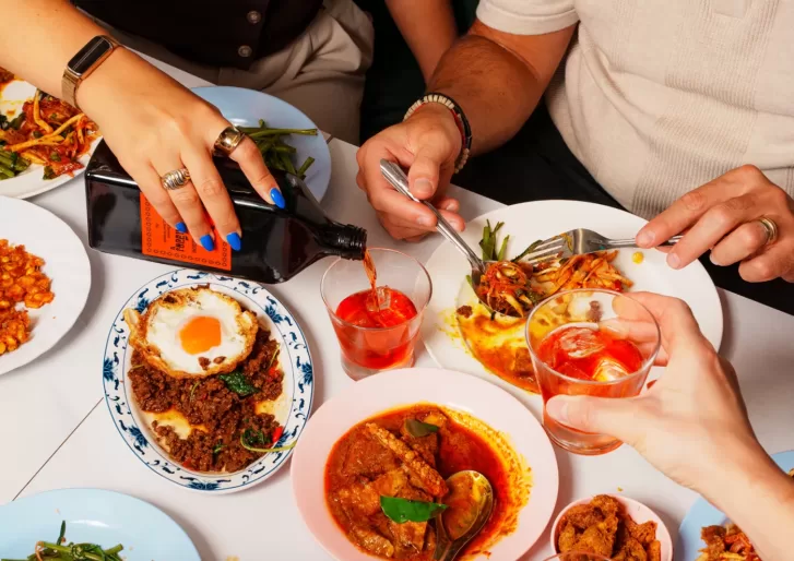 Three people share a colorful meal at a table, serving dishes like curry, a fried egg over minced meat, and salad. One person pours a dark drink into a glass while others toast or eat with forks.